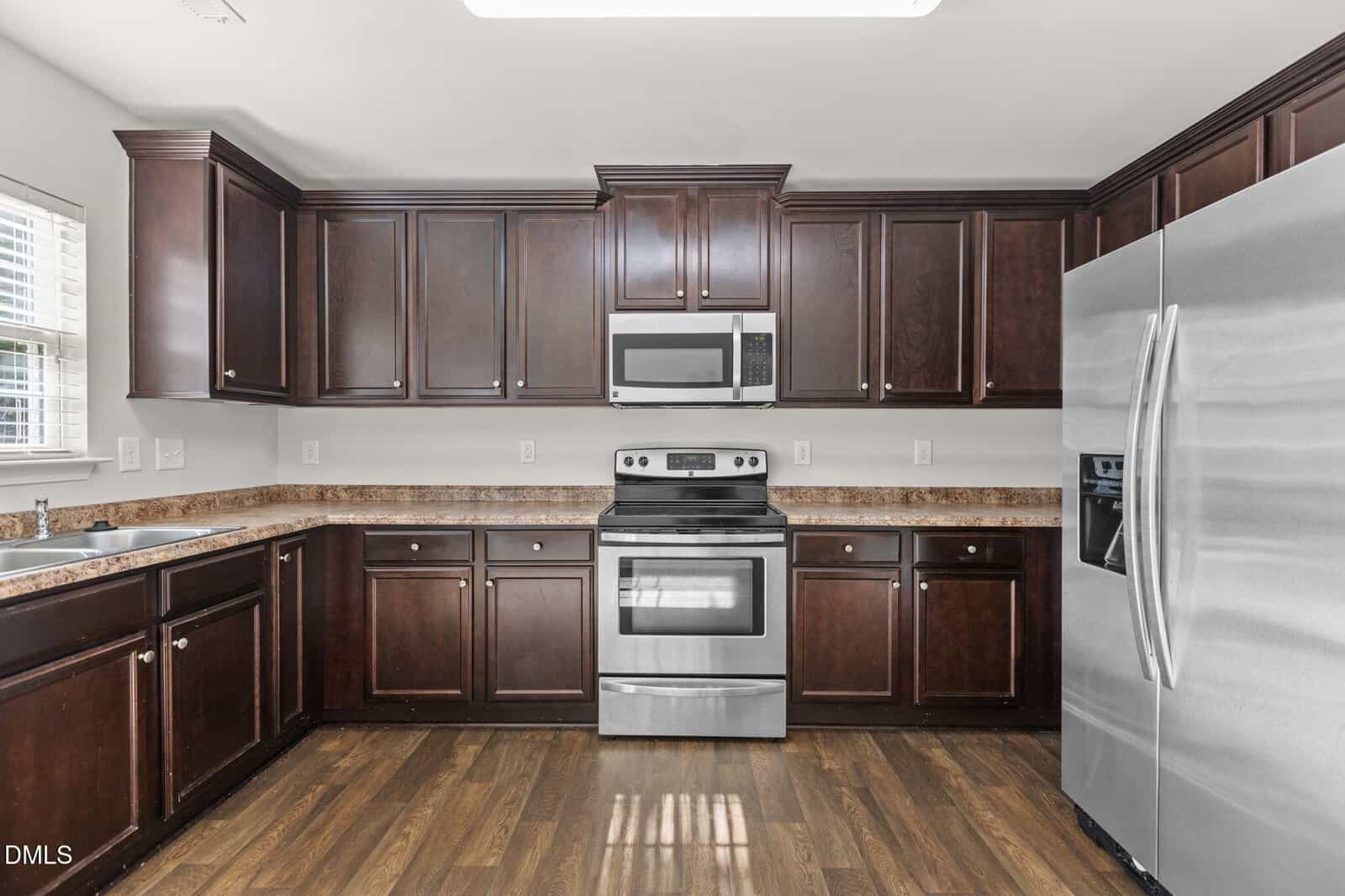 Modern kitchen with dark wood cabinets, stainless steel appliances including an oven, microwave, and refrigerator, brown granite countertops, and wood flooring. A window above the sink lets in natural light.