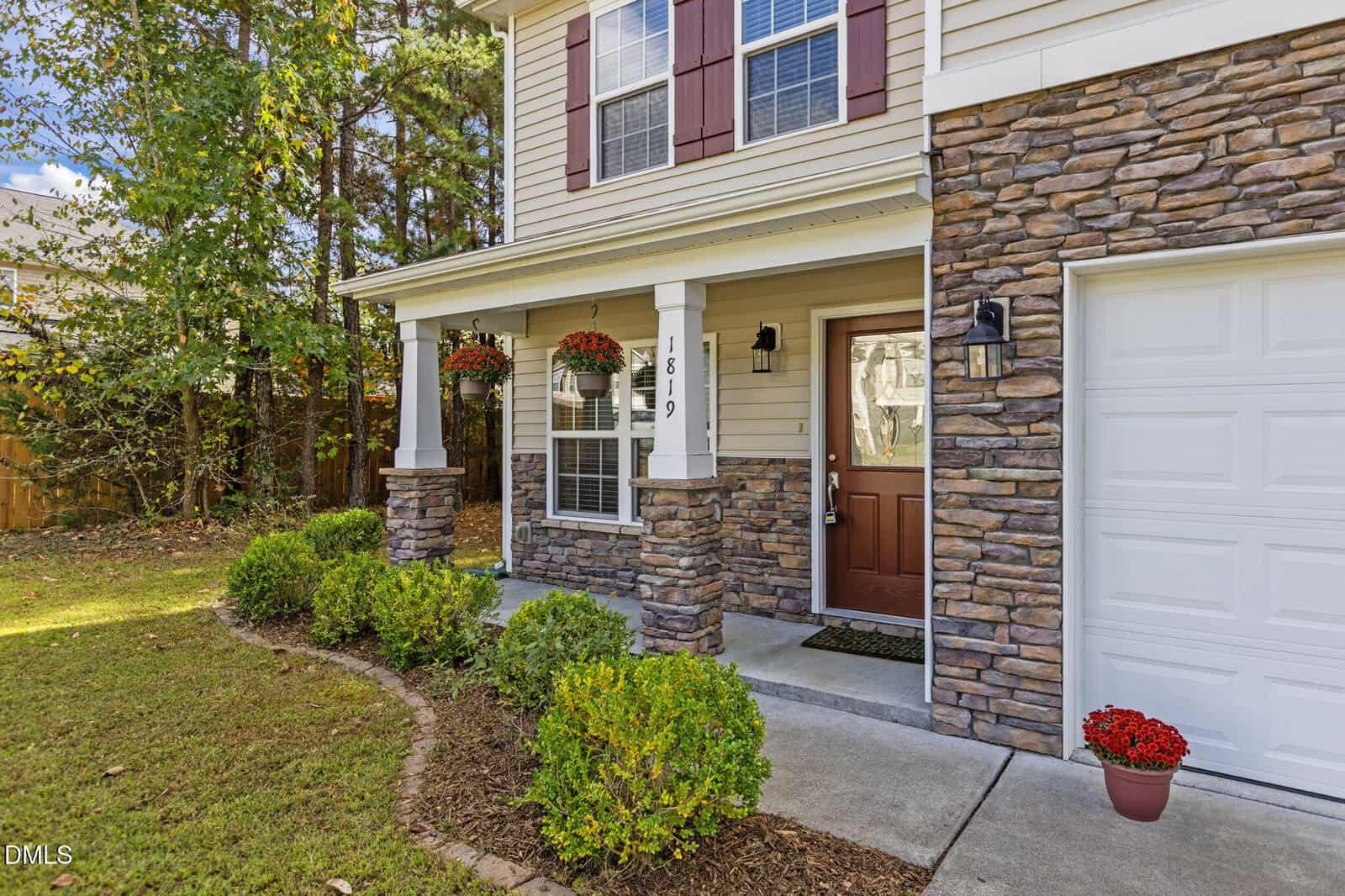 Front exterior of a two-story house with stone accents, a covered porch supported by stone columns, a brown front door, red potted flowers, and shrubs along a curved mulch bed by the sidewalk.