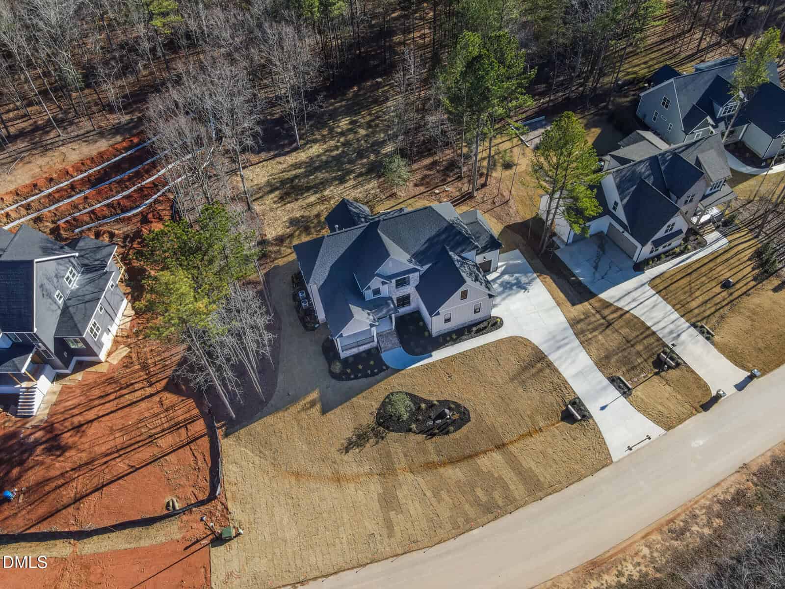 Aerial view of a two-story house with a driveway, lawn, and surrounding trees. Nearby houses and a road are visible, with red soil and landscaping work in progress on one side.