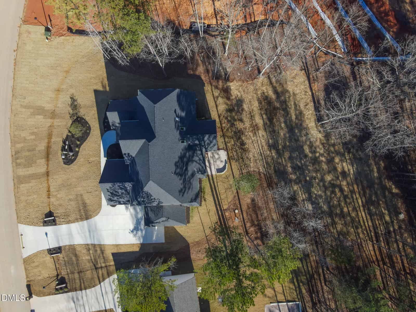 Aerial view of a two-story house with a dark roof, white driveway, and surrounding yard bordered by trees and bare branches, casting shadows on the ground. A concrete patio is visible behind the house.