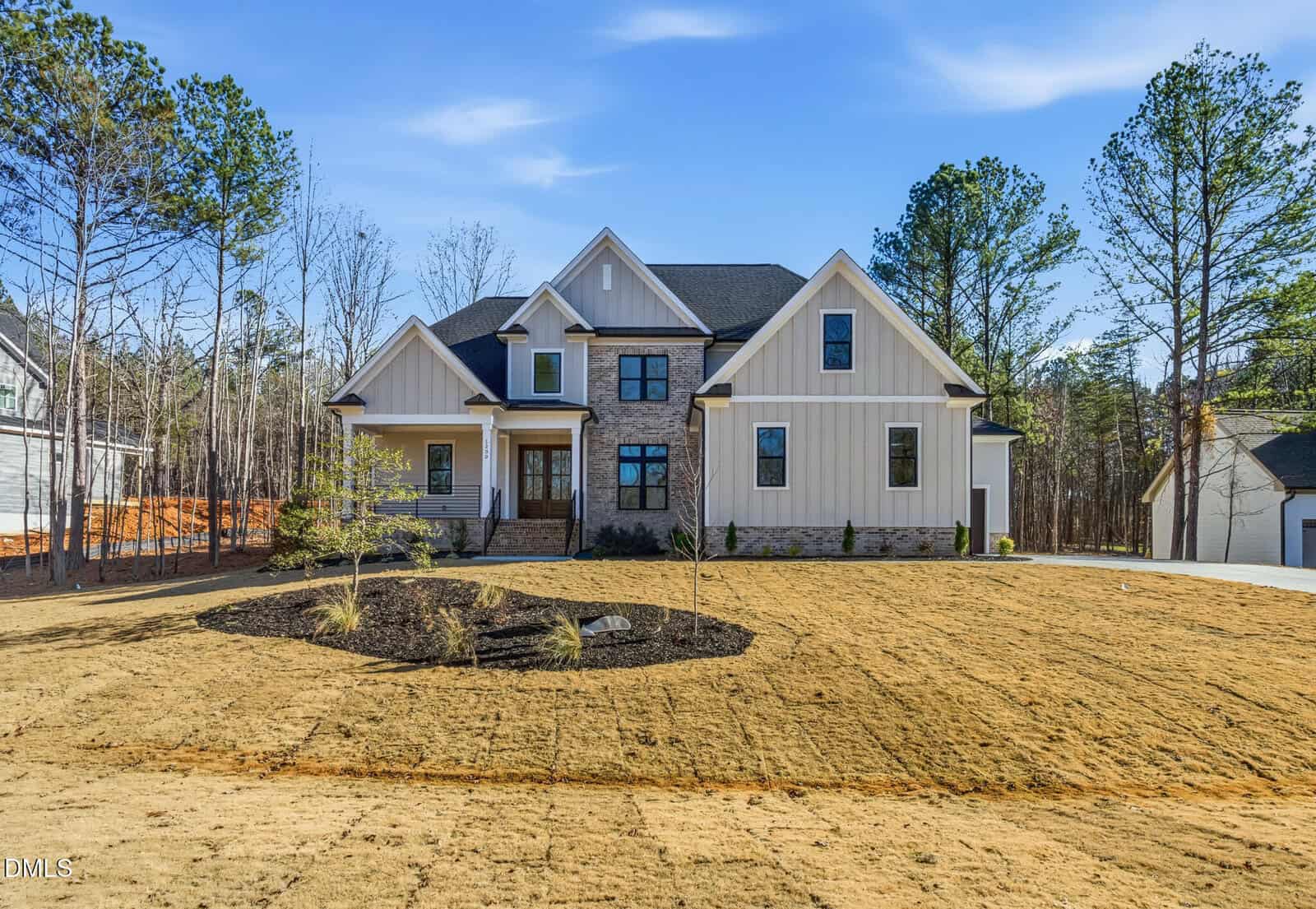A two-story modern farmhouse-style home with stone and light-colored siding, large windows, a covered front porch, and a landscaped front yard with new grass and young trees. Tall trees line the property in the background.