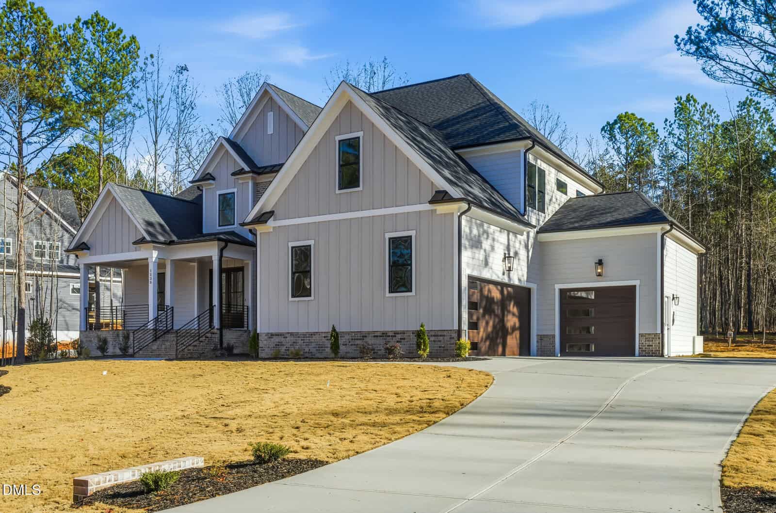 A modern, two-story suburban house with light-colored siding, dark roof, attached garage, and a spacious curved driveway. The front yard has sparse grass and minimal landscaping; trees are visible in the background.