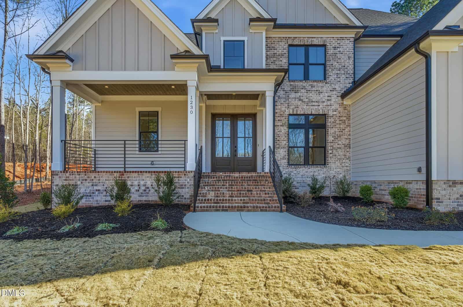 Two-story modern house with brick and siding exterior, covered front porch, double front doors, black railings, and landscaped yard with small shrubs and fresh sod.