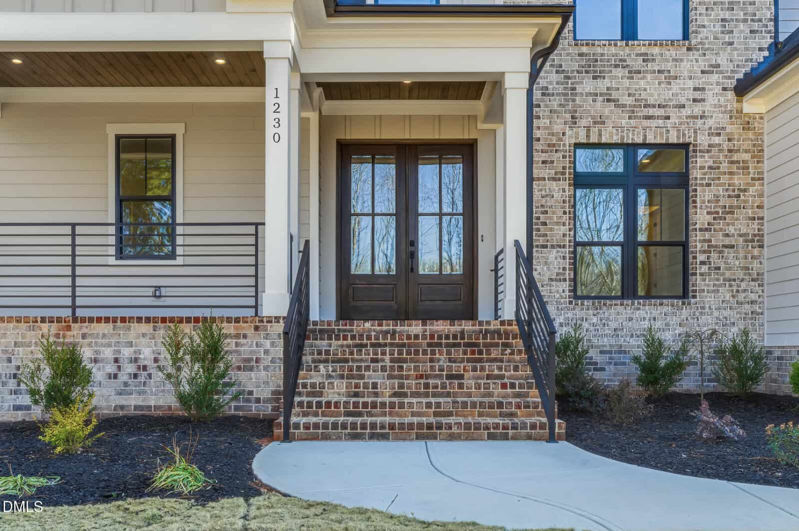 The image shows the front entrance of a house with a brick staircase leading to double wooden doors, flanked by windows and simple landscaping with small bushes. The house number 1230 is visible by the doorway.