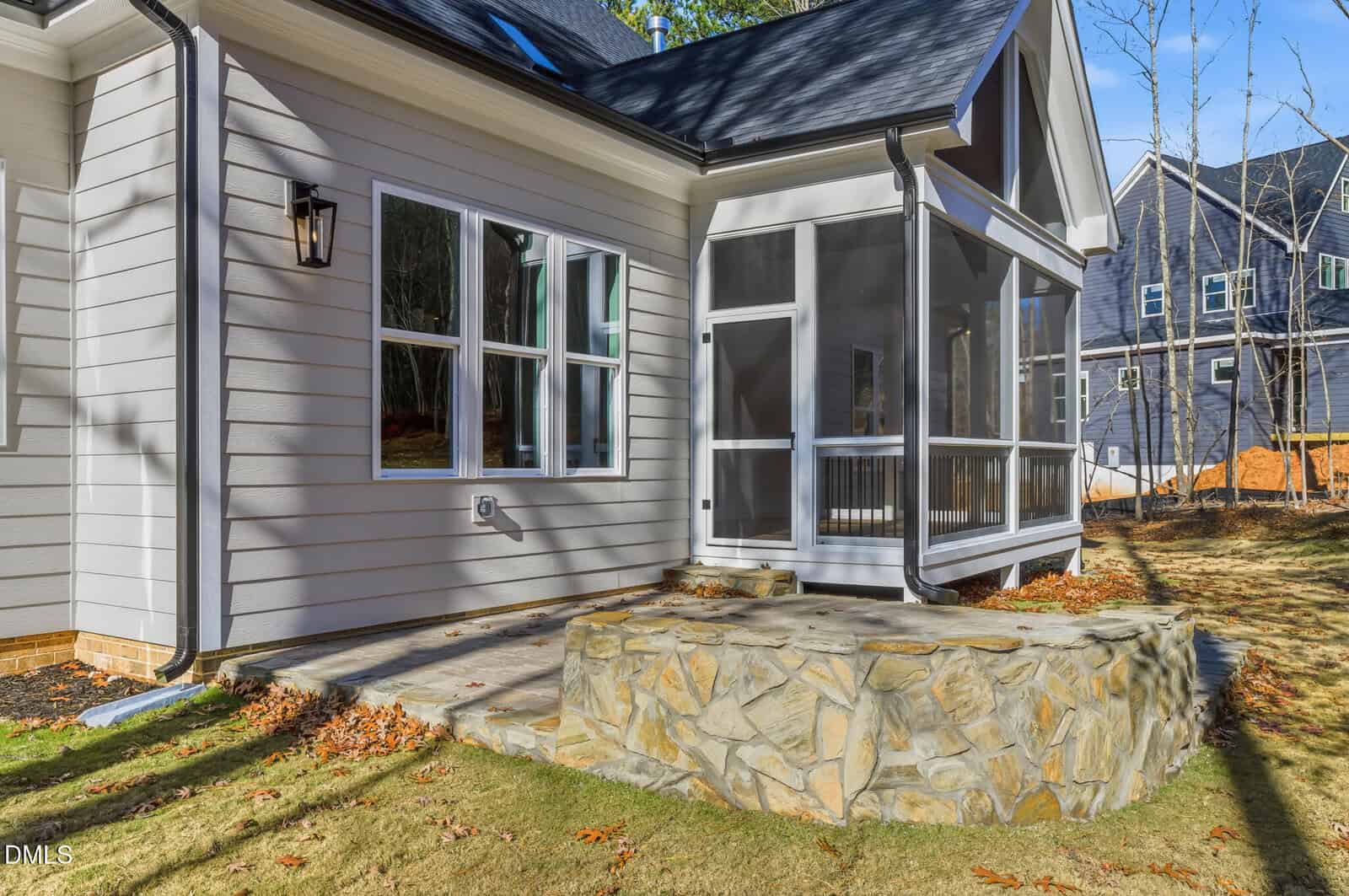 Side view of a house with light siding, a screened porch, a stone patio area, outdoor light fixture, and grass yard. Another house is visible in the background with trees and clear skies.