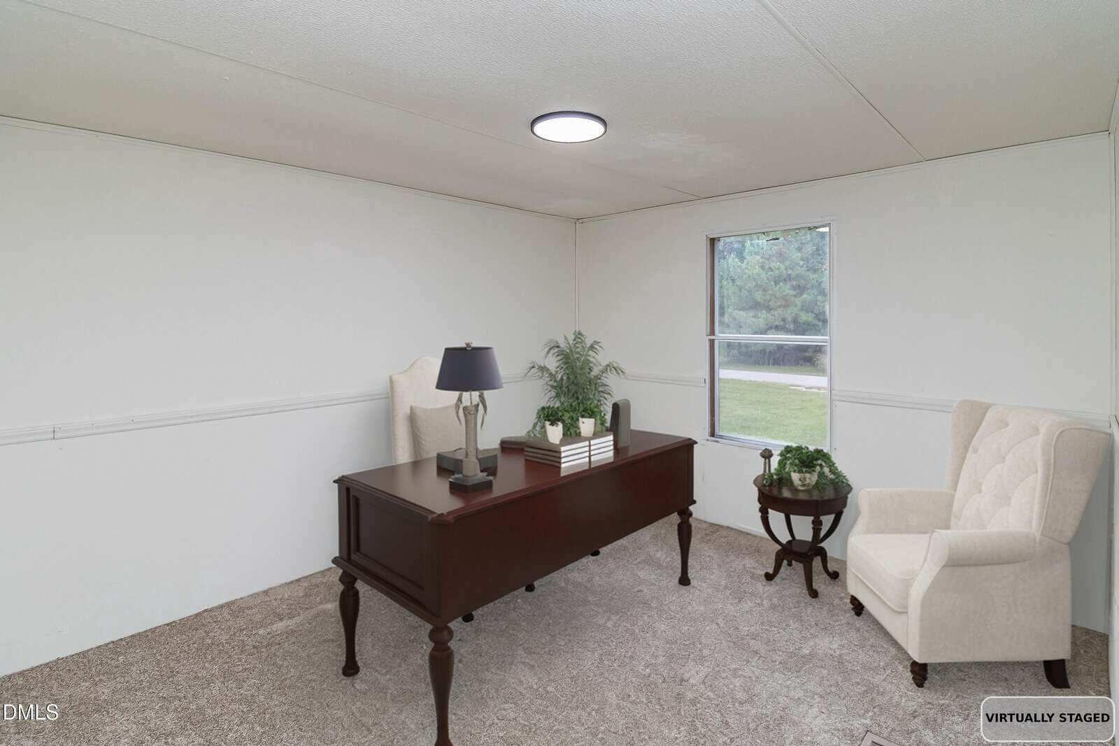 A small, simply decorated office with a dark wood desk, a lamp, plant, and chair; another armchair and a small round side table are by the window. The room has beige carpet and white walls.