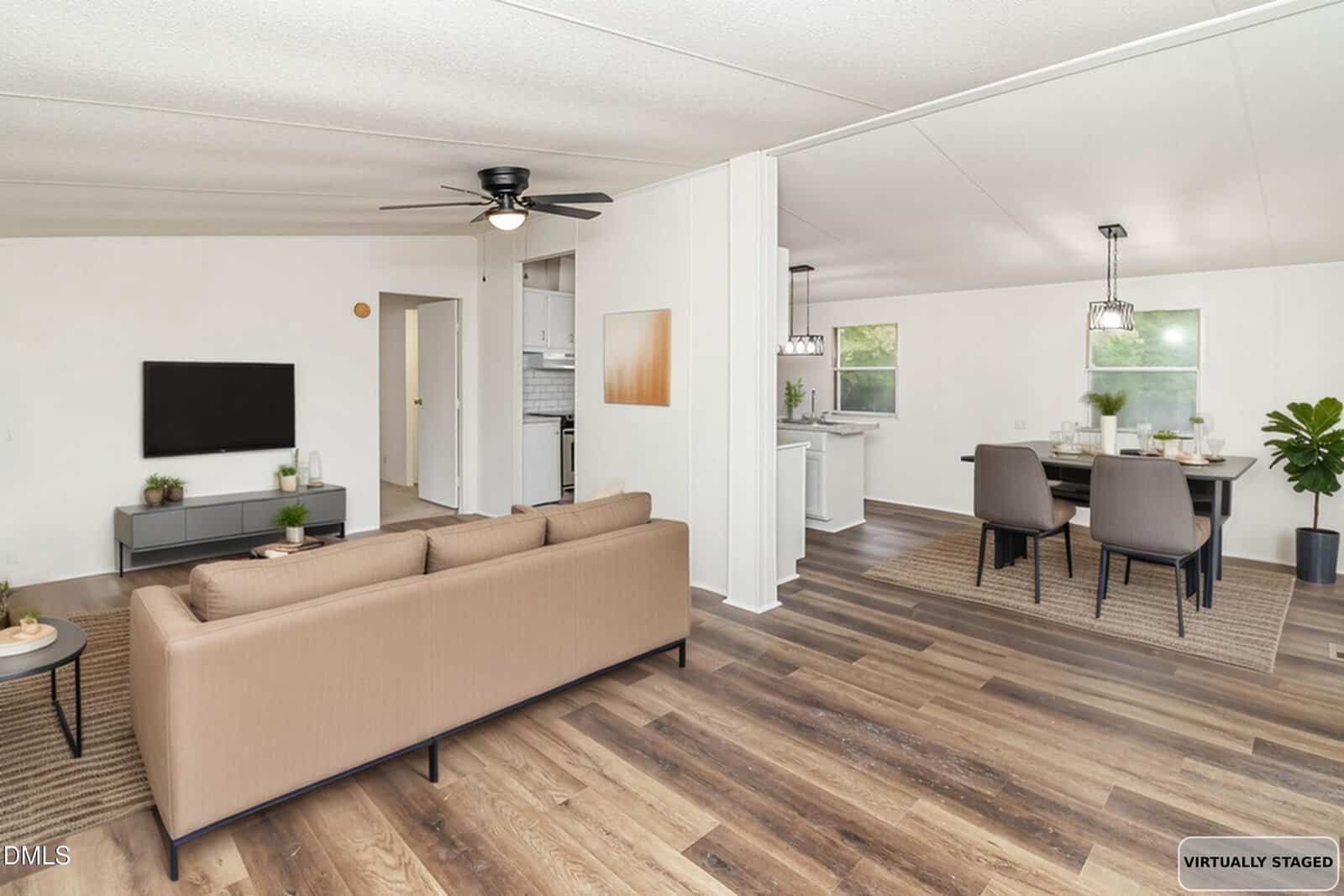 A bright, open living and dining area with wood flooring, a beige sofa facing a wall-mounted TV, a dining table with six chairs, and large windows letting in natural light. The space is virtually staged.
