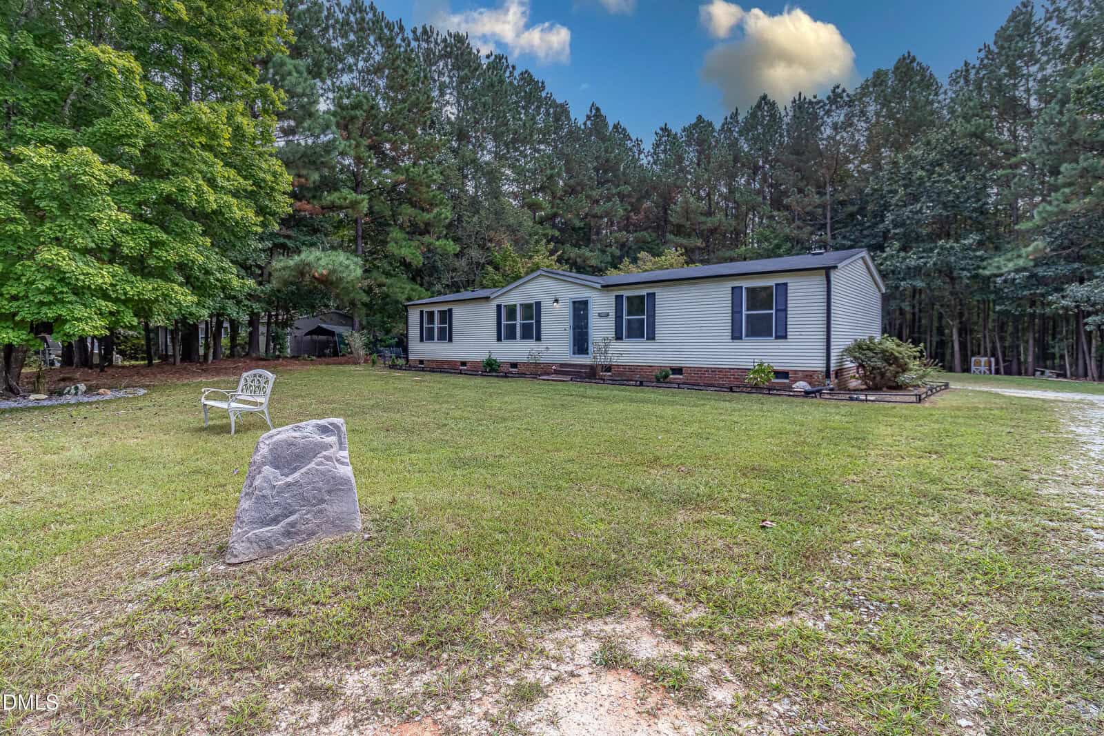 A single-story white house with blue shutters sits on a large grassy yard bordered by a forest of tall pine trees. A white bench and a large stone are in the foreground near the driveway.