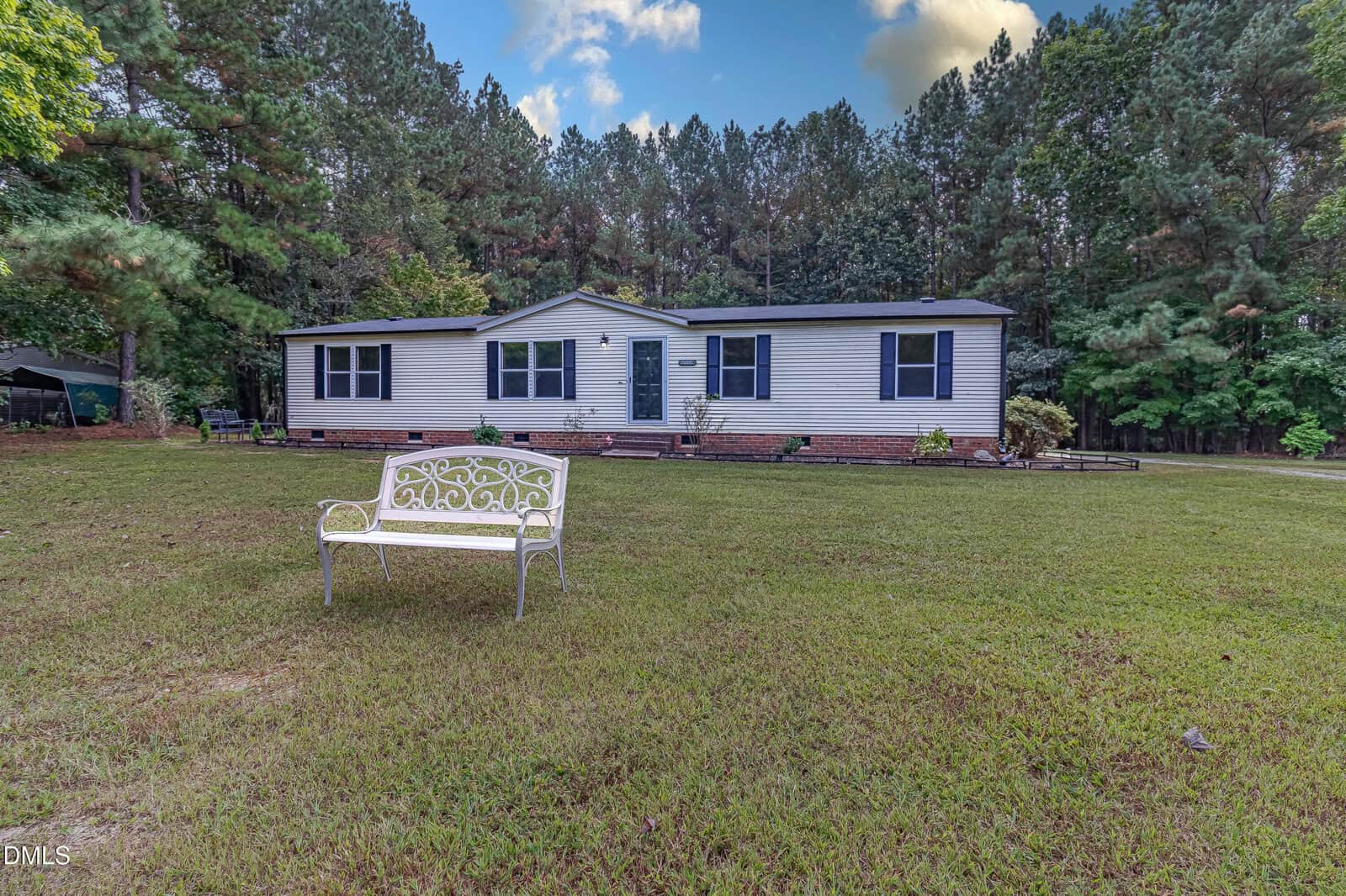 A single-story white house with dark shutters sits on a large grassy lawn at 110 Castlebury Lane, Franklinton, featuring a white decorative bench in front and surrounded by tall trees with dense green foliage under a partly cloudy sky.
