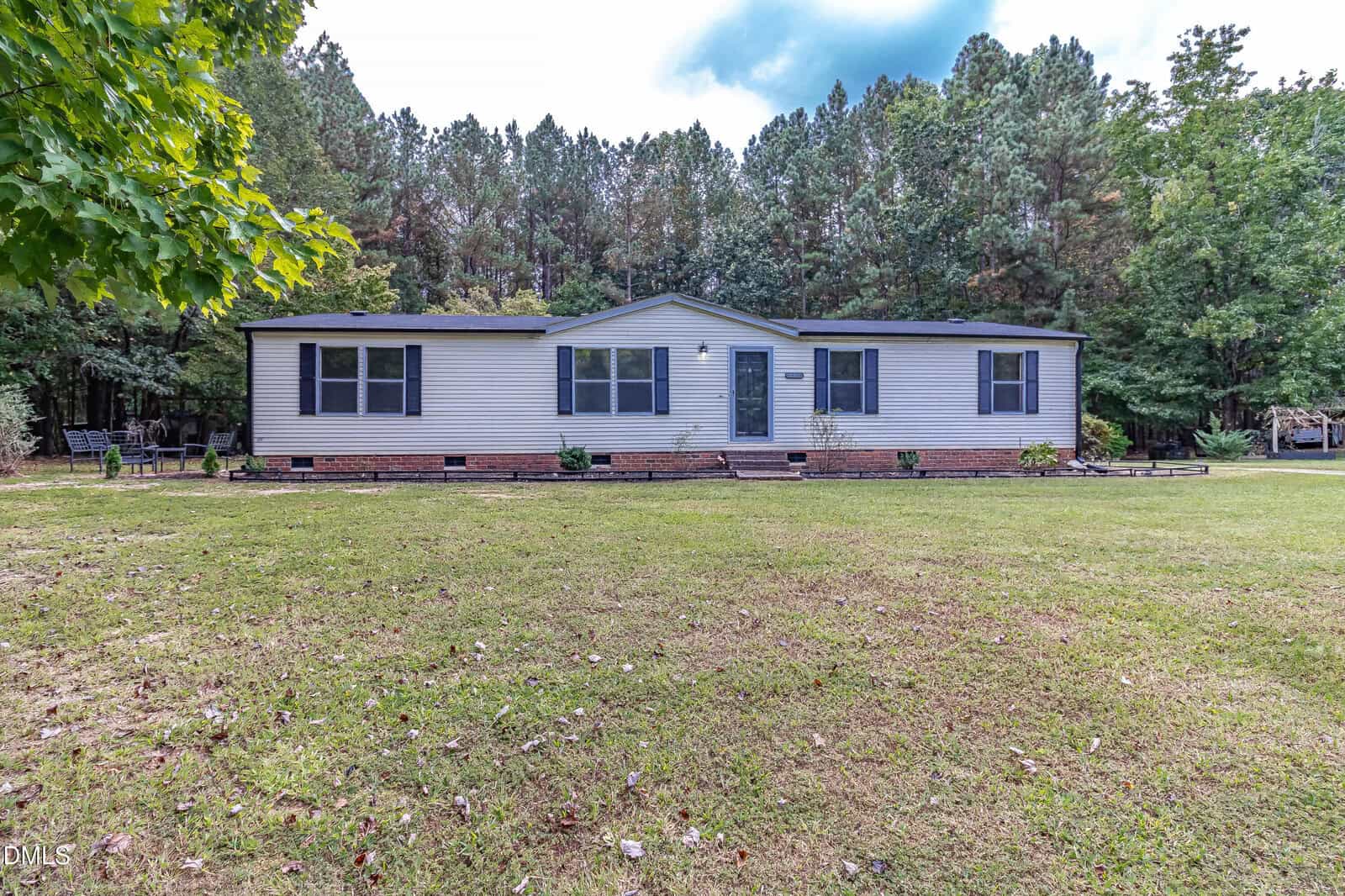 A single-story, beige manufactured home with dark shutters and a central front door, set on a grassy lawn with trees in the background.