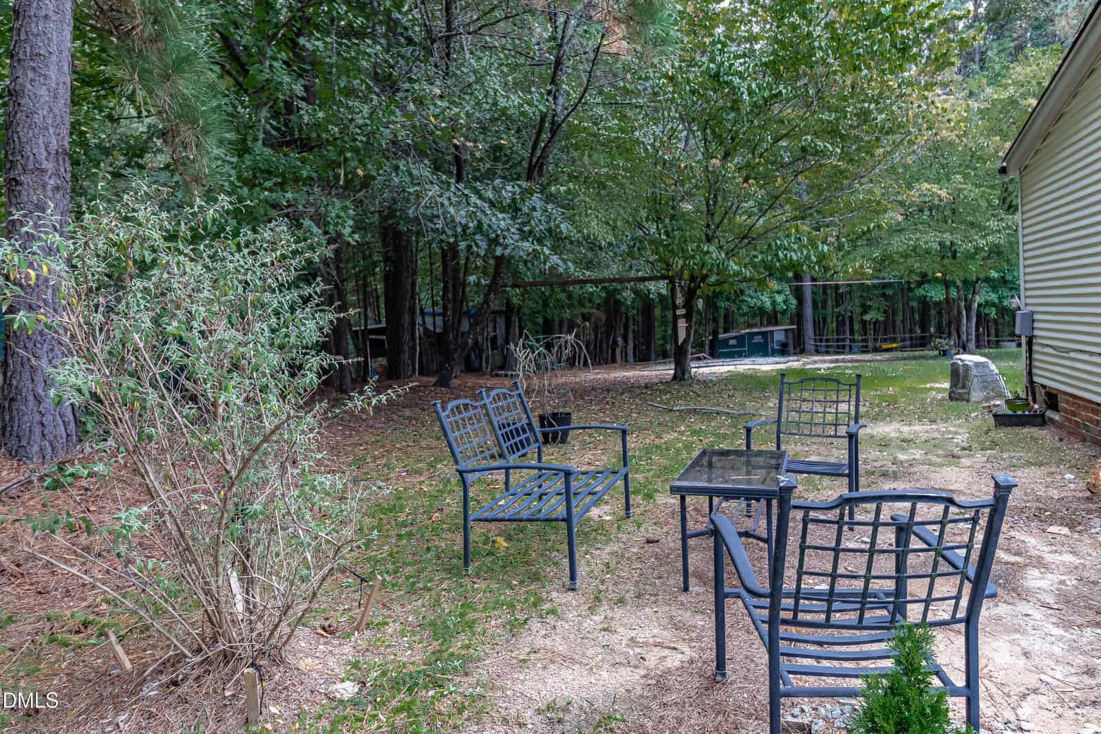 Four metal chairs and a small glass table are arranged on a patch of dirt and pine needles in a shaded backyard, surrounded by trees and greenery next to a house with yellow siding.