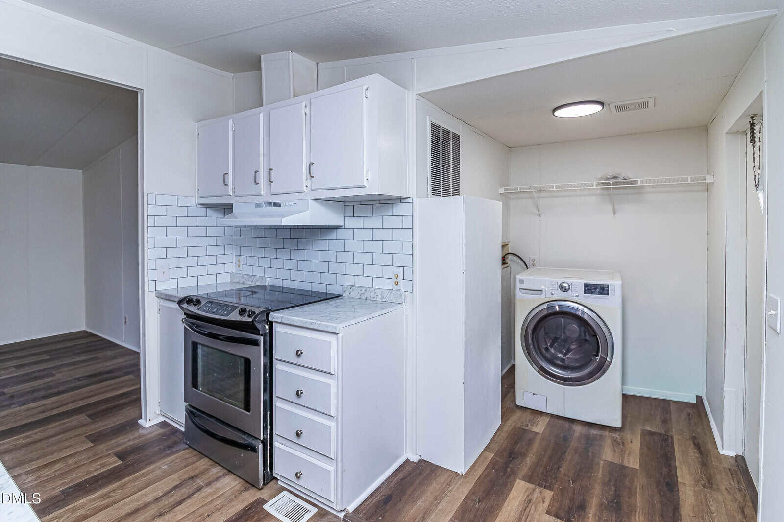 A small kitchen with a black stove, white cabinets, white subway tile backsplash, and marble countertop is next to a laundry area with a front-loading washer and open shelving. The floor is dark wood.