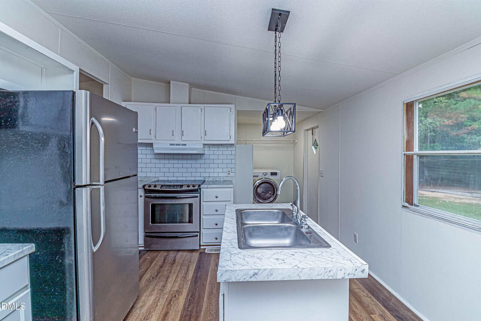 A modern kitchen with white cabinets, stainless steel appliances, a marble island with a double sink, a hanging light fixture, and a view of a laundry area in the background. Large window lets in natural light.