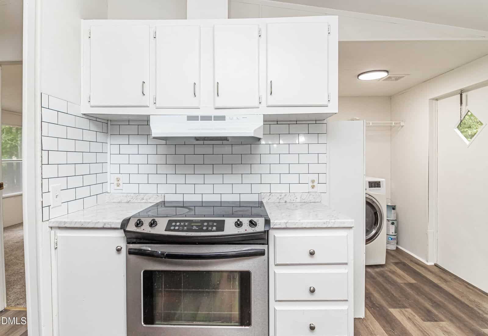 Modern kitchen with white cabinets, white subway tile backsplash, marble countertop, stainless steel oven, and stovetop. Laundry area with washer and dryer is visible in the background. Wood-style flooring throughout.