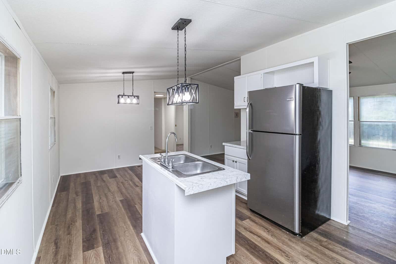Modern kitchen with white cabinets, marble countertop island with sink, stainless steel refrigerator, and wood flooring, open to a bright dining and living area with hanging light fixtures.