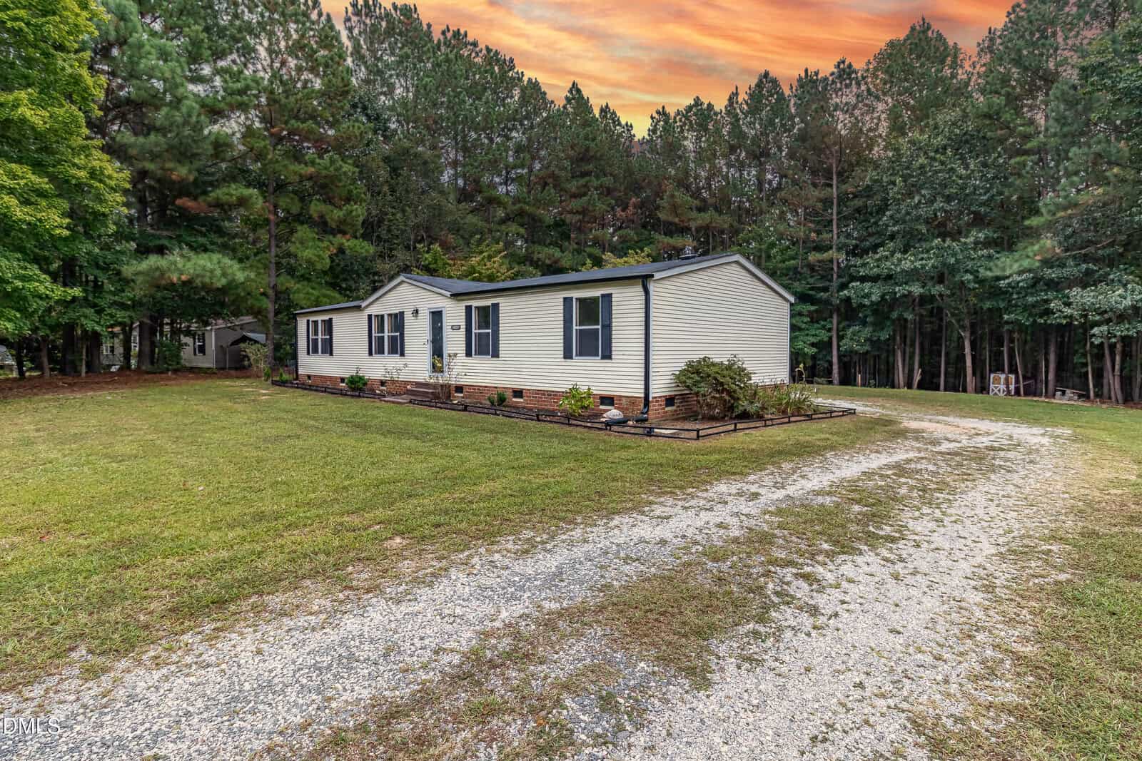 A single-story beige house with blue shutters sits on a spacious grassy lot, bordered by tall trees. A gravel driveway curves in front of the house under a vibrant orange and pink sunset sky.