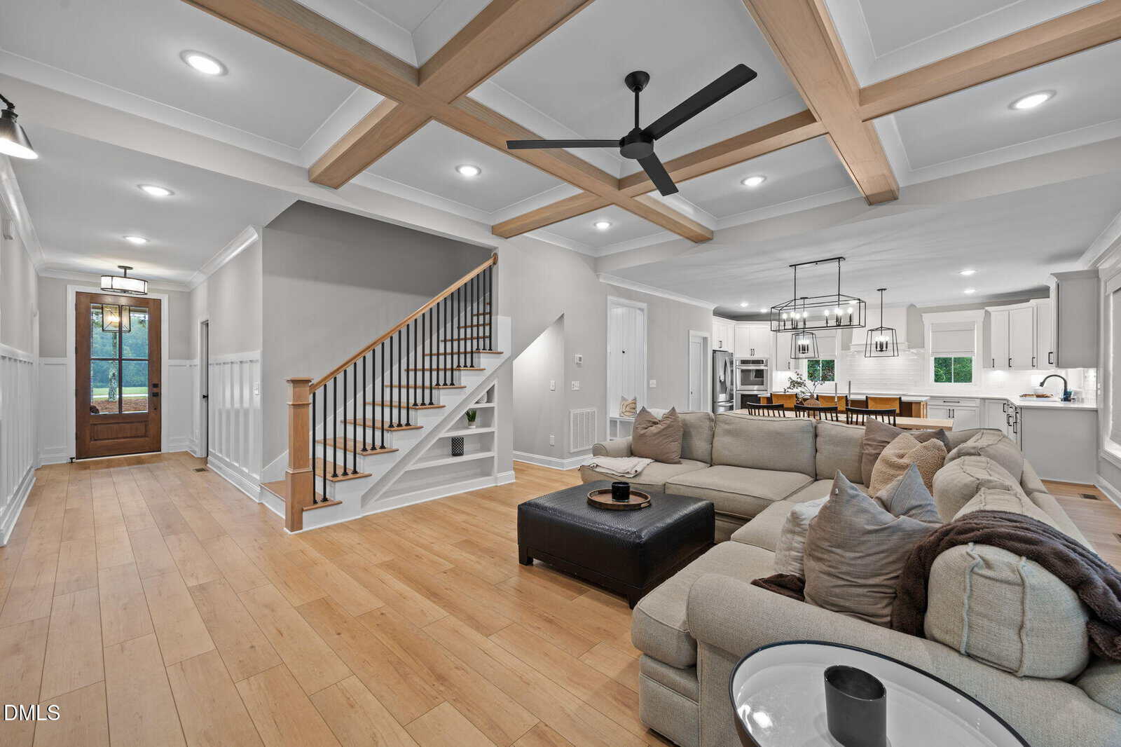 Spacious living room with light wood floors, beige sectional sofa, black ottoman, and exposed ceiling beams. Open floor plan includes kitchen and dining area. Staircase and wooden front door are visible in the background.