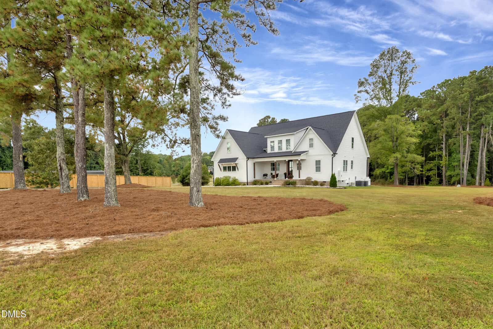 A large white two-story house with a covered front porch sits on a spacious grassy lot, surrounded by tall pine trees under a blue sky with scattered clouds.