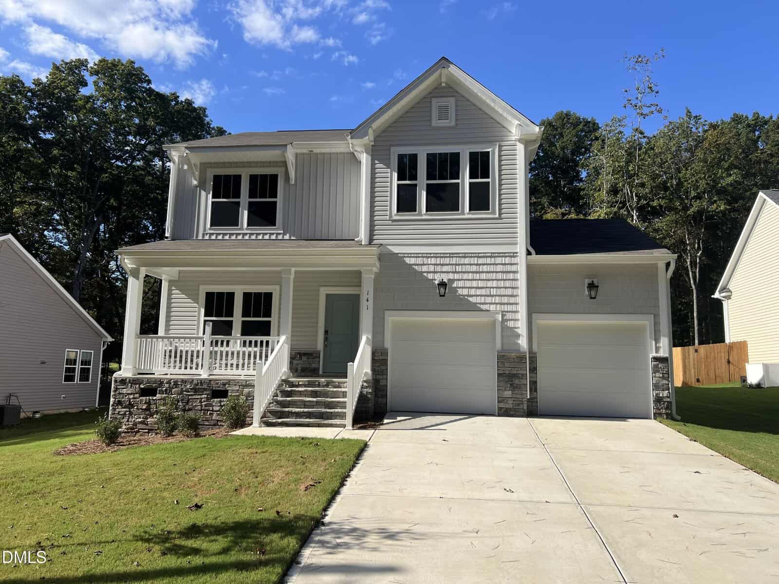 Two-story gray house with white trim, a covered front porch, stone accents, double garage, and concrete driveway located at 141 Oswego Drive 2508 in Louisburg. The yard is grassy with a few trees, and the sky is clear and blue.