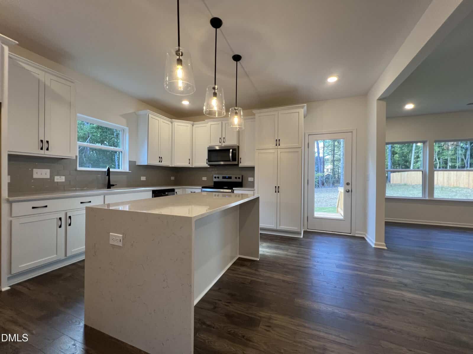 Modern kitchen with white cabinets, stainless steel appliances, a central island with pendant lights, and dark wood flooring. Large windows and a glass door let in natural light; open space leads to another room.