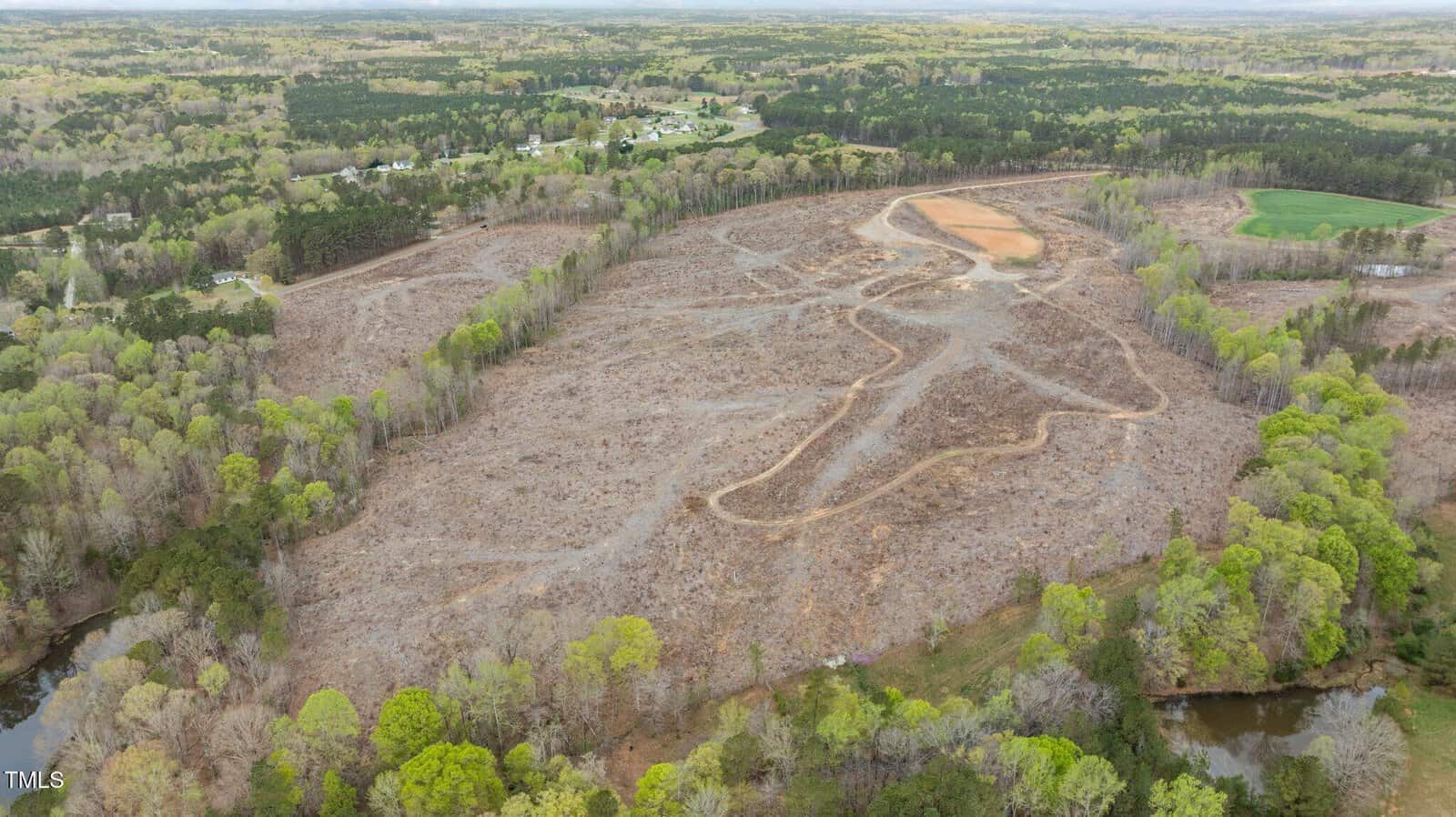 A large, cleared plot of land with visible dirt paths and minimal vegetation, surrounded by trees and forested areas; residential buildings are visible in the background.