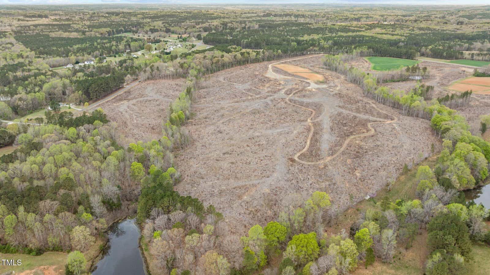 Aerial view of a large, cleared area of land with a winding dirt path, surrounded by patches of forest, a pond in the foreground, and scattered houses and fields in the distance.