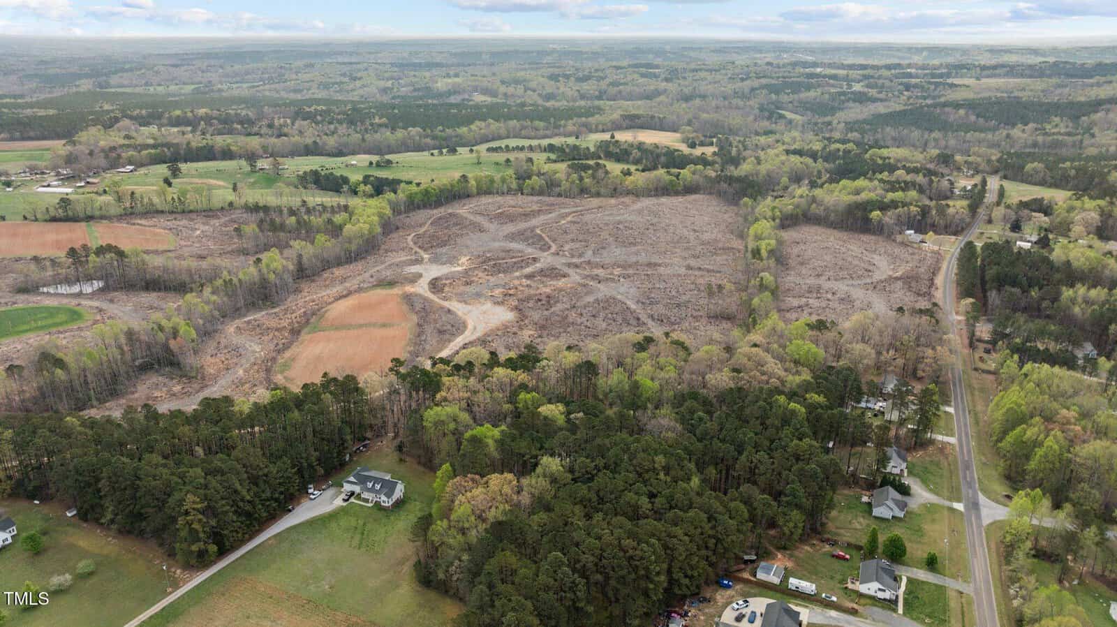 Aerial view of a rural area with a large, clear-cut section of land surrounded by trees, open fields, scattered houses, and a narrow road running along the right side of the image under a cloudy sky.