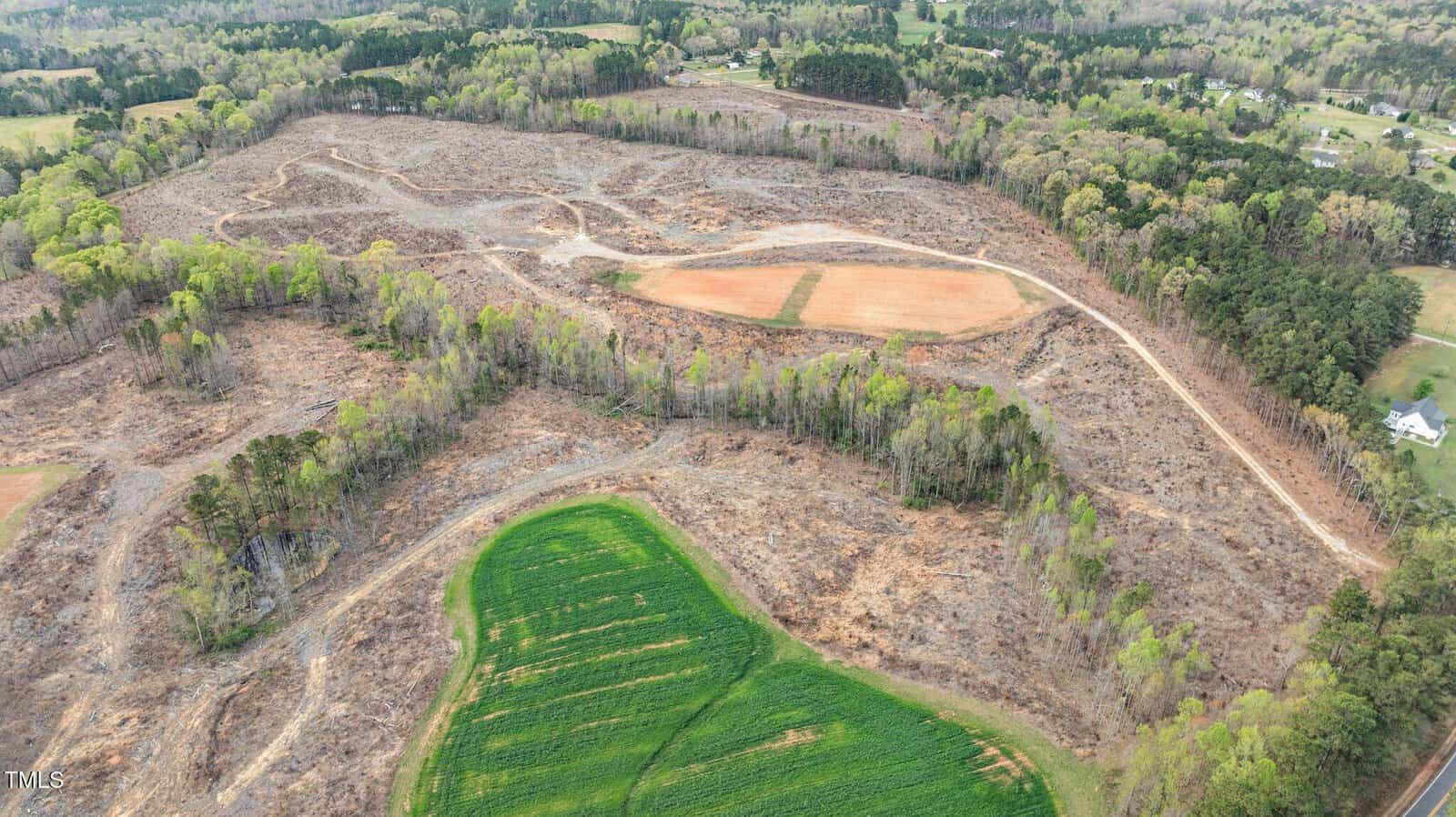 Aerial view of a large, partially cleared area of land with some remaining trees, dirt paths, and green fields. Surrounding areas are wooded with a few scattered houses visible in the distance.