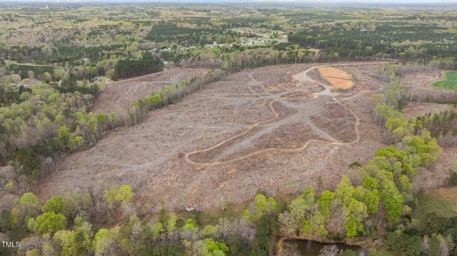 Aerial view of a large, cleared area of land with dirt paths winding through it, surrounded by dense green forest and a few distant houses. The sky is overcast, and a small pond is visible in the foreground.