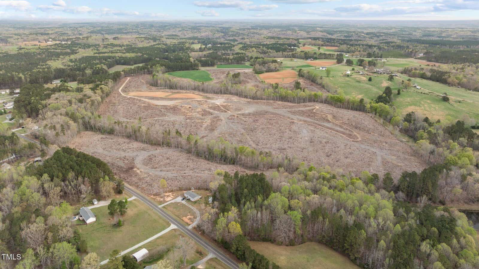 Aerial view of a large, cleared plot of land with winding dirt paths, surrounded by green forests, scattered fields, and residential houses under a partly cloudy sky.