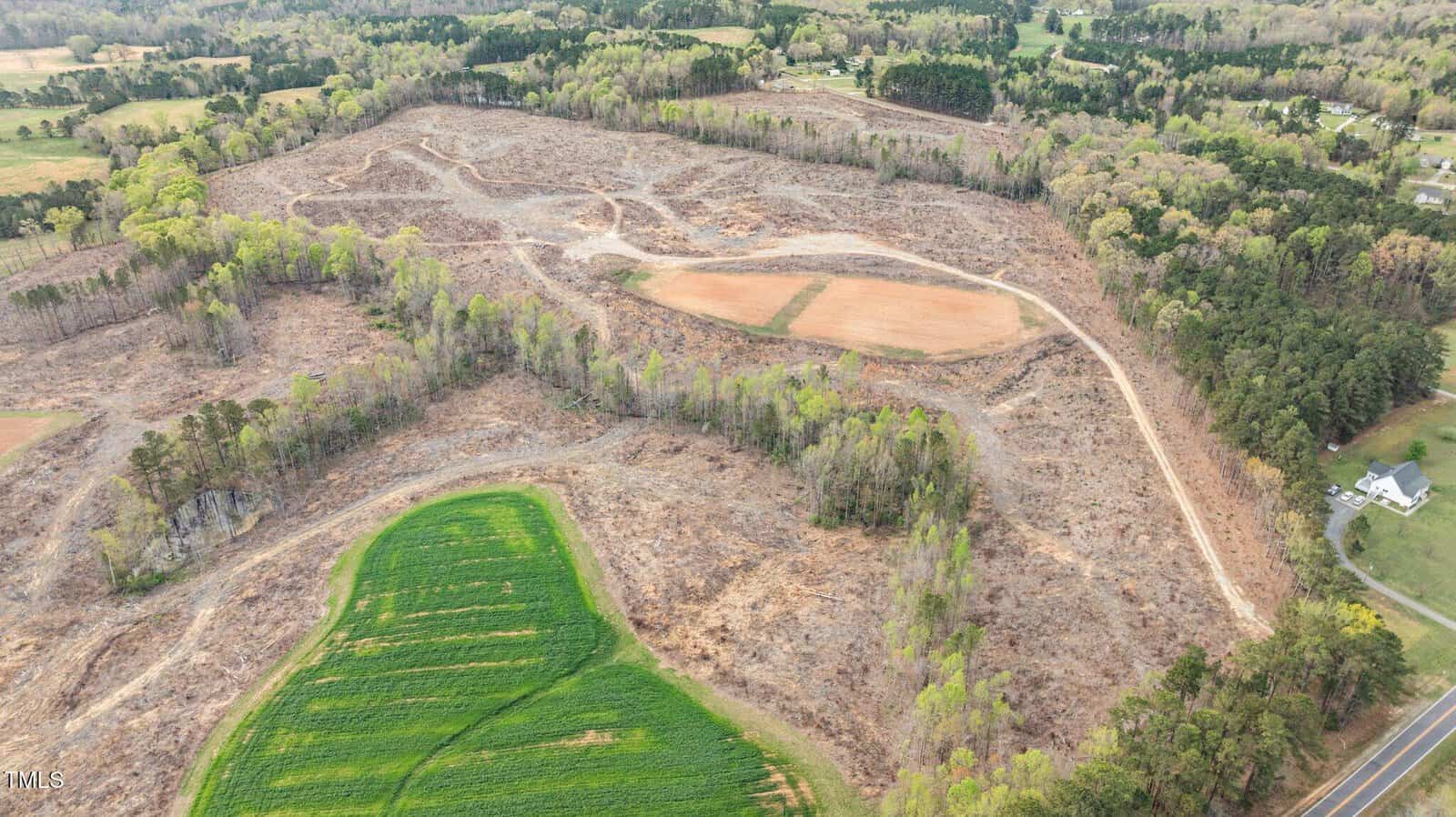 Aerial view of a large, partially cleared area of land with patches of remaining trees, a dirt road winding through, and some farmland adjacent to a paved road and houses in the lower right corner.