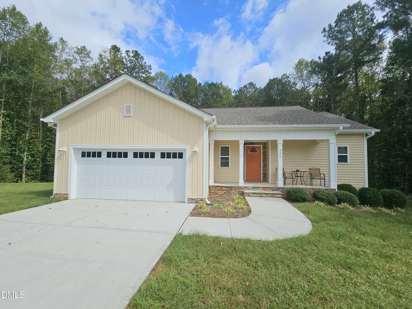 Single-story beige house with a two-car garage, front porch, two chairs and a table, concrete driveway, and green lawn. Surrounded by trees and partly cloudy blue sky.