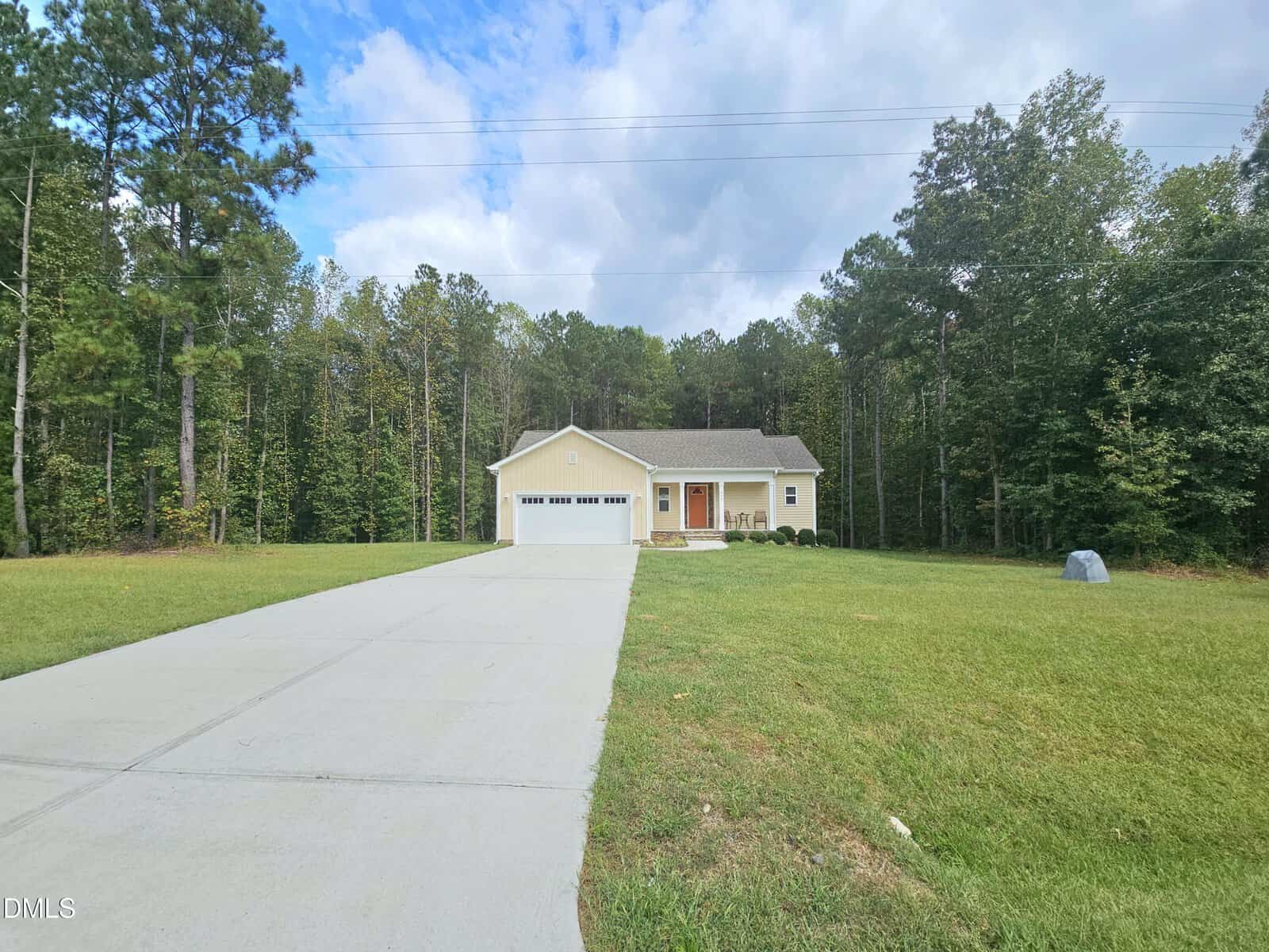 Single-story house with a two-car garage, tan siding, and a covered front porch, situated at the end of a long driveway, surrounded by grass and tall trees under a partly cloudy sky.
