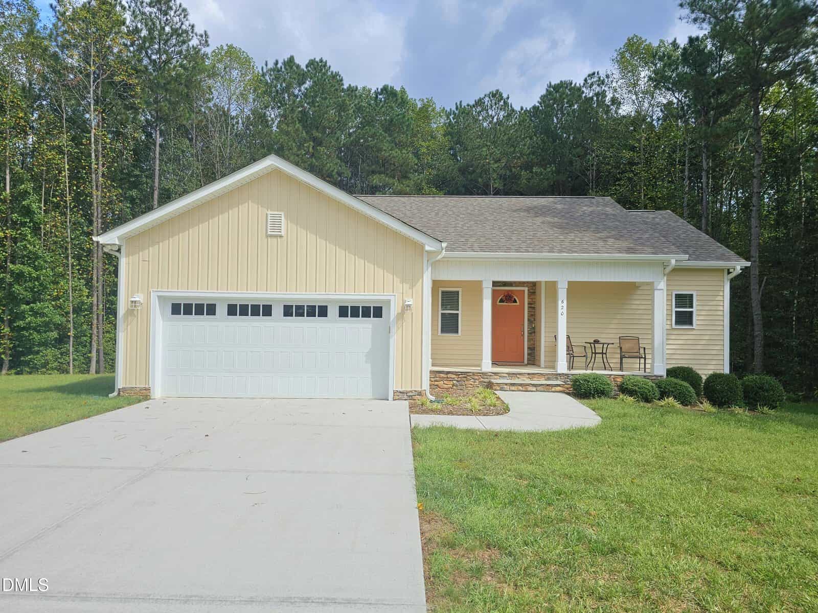 Charming single-story beige house located at 620 Raymond Tharrington Road in Louisburg, featuring a two-car garage, an inviting orange front door, a small covered porch with two chairs, concrete driveway, and a grassy lawn set against a backdrop of tall trees.