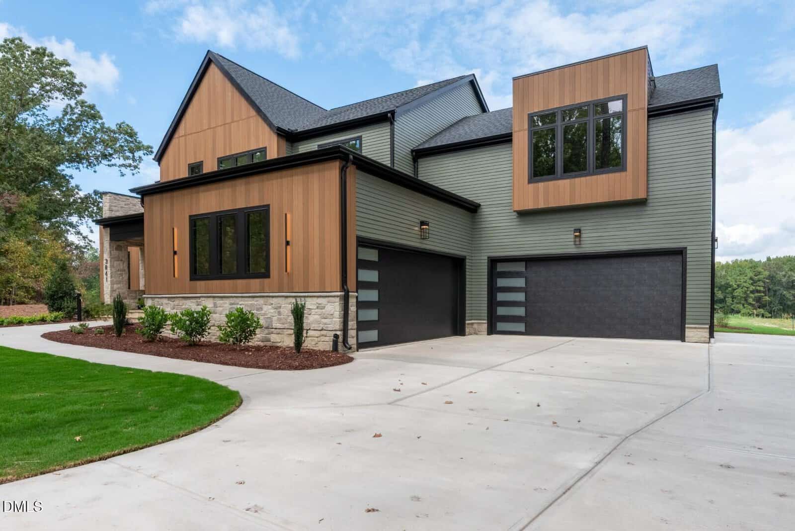 A modern two-story house with wood and sage green siding, black framed windows, three-car garage with frosted glass doors, concrete driveway, and landscaped yard with grass and mulch.