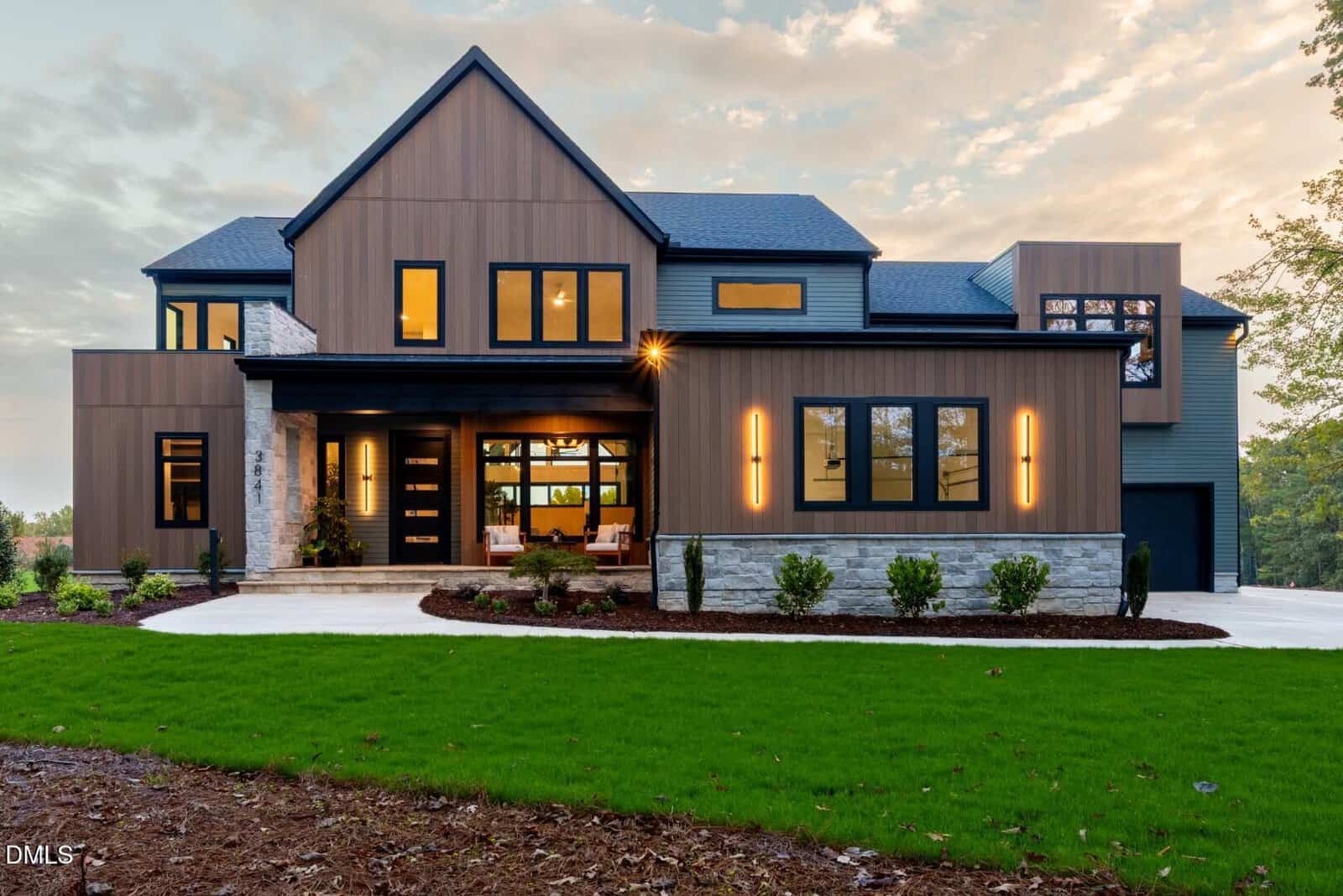 Contemporary two-story house with wood paneling, large windows, stone accents, and modern exterior lighting. Well-maintained green lawn and paved driveway in front, with trees in the background.