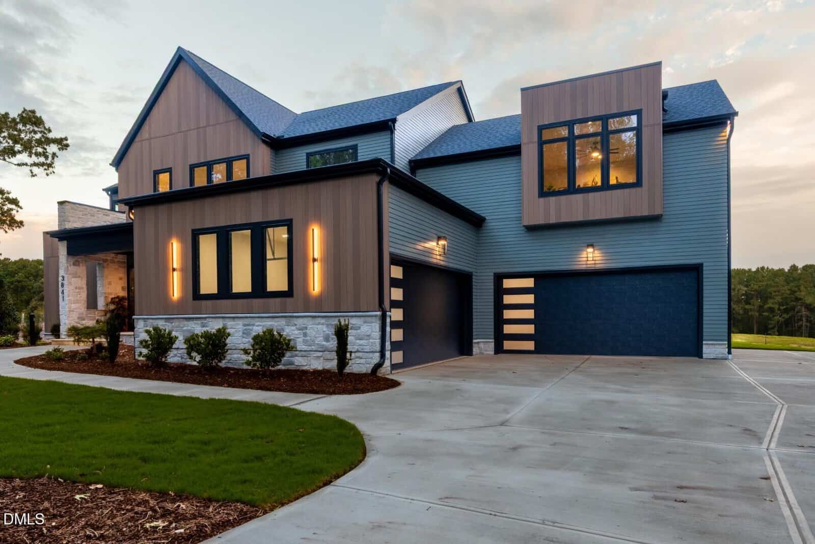 Modern two-story house with blue and brown exterior, large windows, and stone accents. The driveway is wide and curves toward the attached three-car garage. The yard has fresh mulch and neatly trimmed grass.