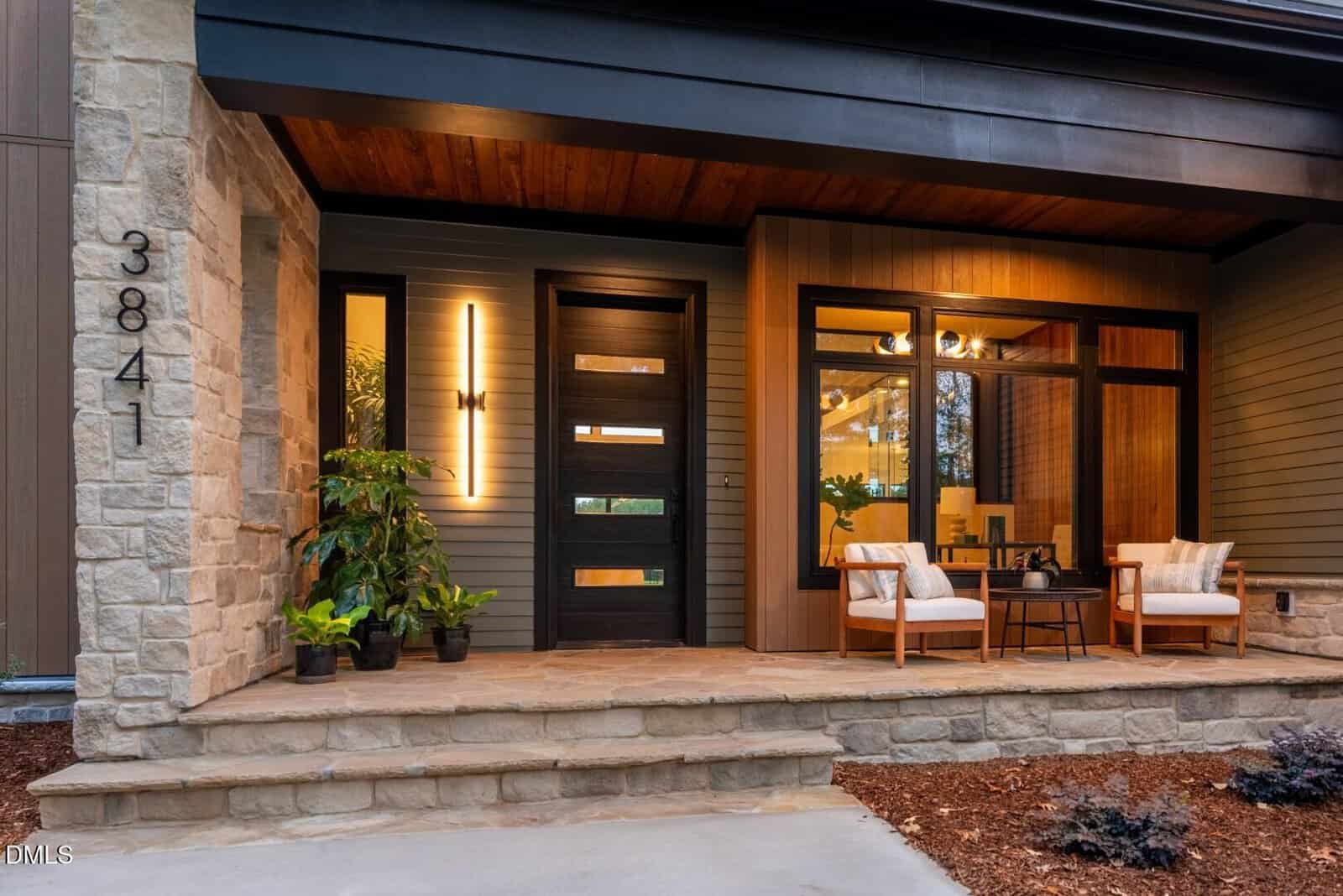Modern house front entry with stone and wood accents, glass-paneled door, house number 3841, two white cushioned chairs with a small table on the porch, potted plants, and warm lighting.