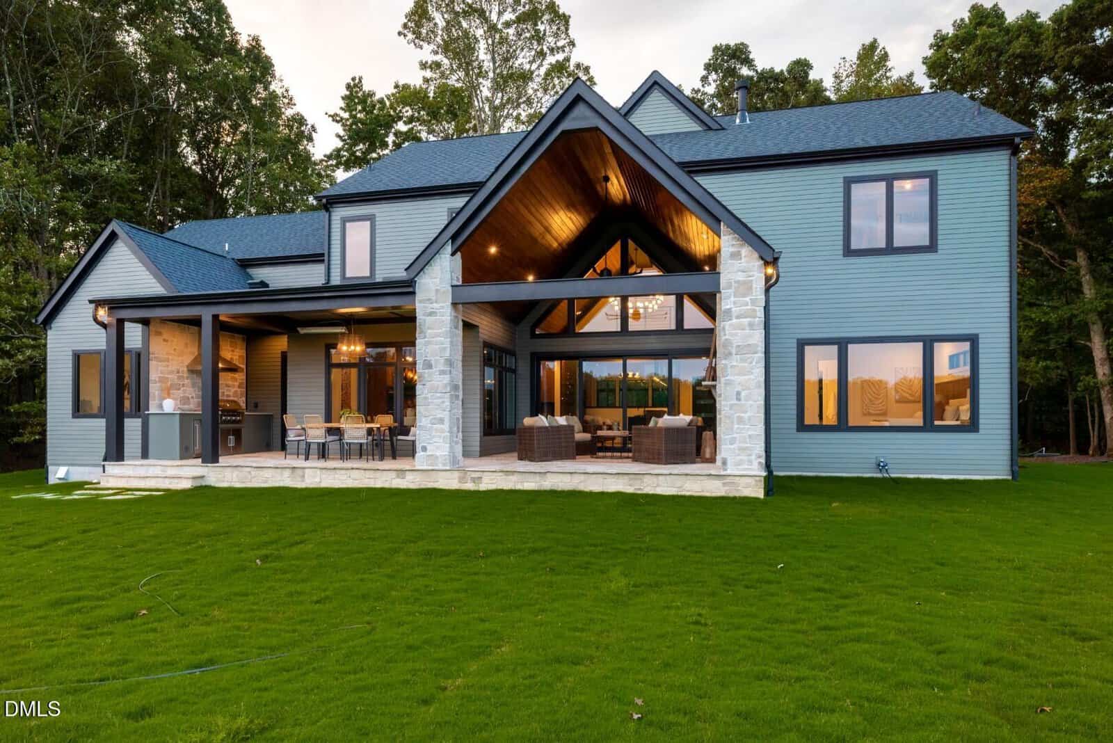 A modern two-story house with blue siding, large windows, a covered outdoor patio, and a manicured green lawn, surrounded by trees in the background.