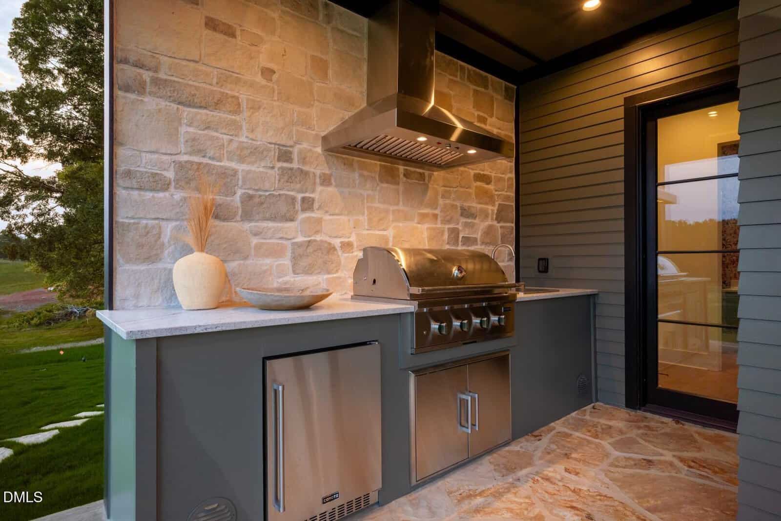 An outdoor kitchen with a stainless steel grill, range hood, mini fridge, and cabinets set against a stone wall. A decorative vase and shallow bowl sit on the countertop. Stone tile flooring is visible.