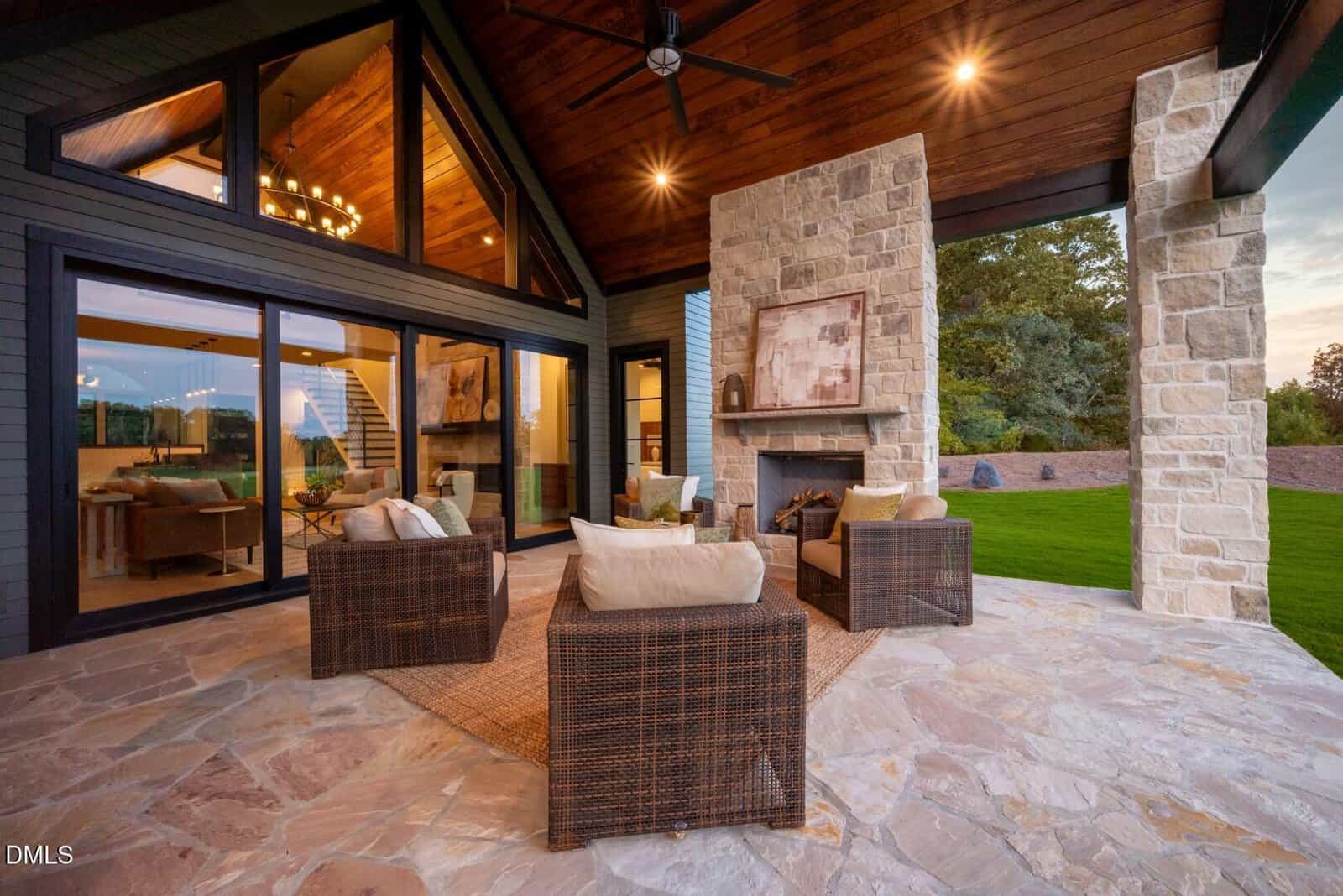A covered patio with stone flooring features four wicker chairs around a stone fireplace. Large glass doors reveal a modern interior with a dining area and staircase. Lush green lawn and trees are visible outside.