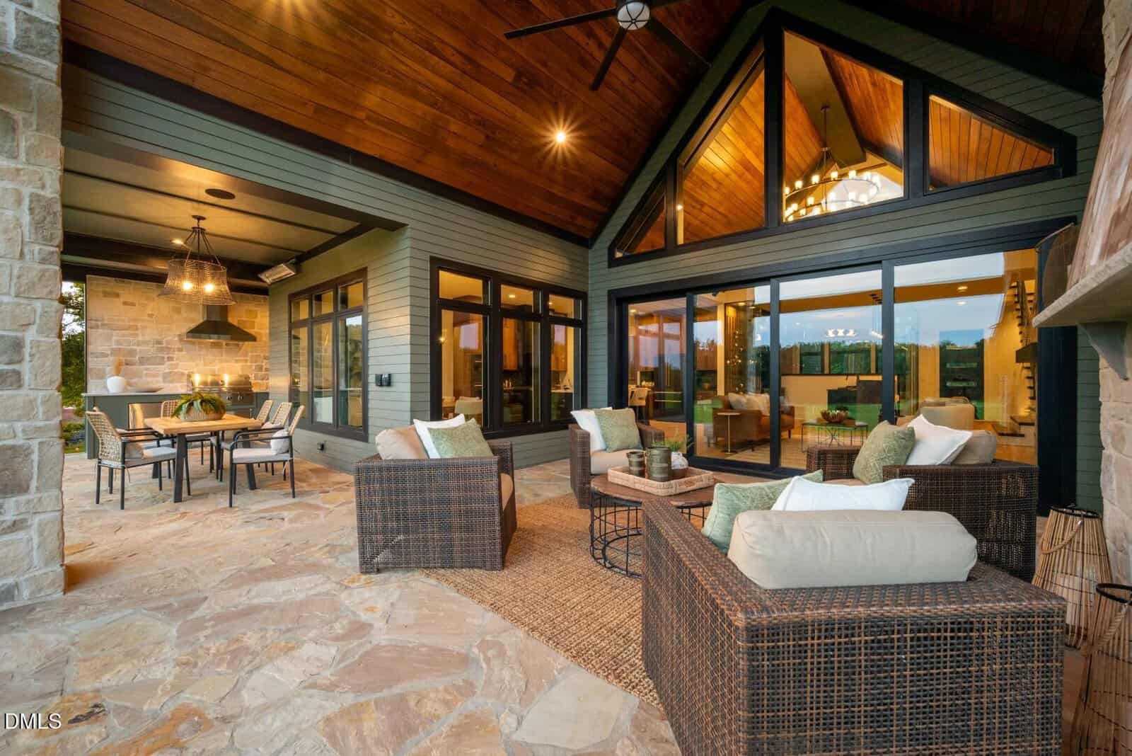 A covered outdoor patio features wicker furniture with light cushions, a dining table with chairs, stone flooring, and large windows and glass doors leading into a modern home with wood-paneled ceilings and warm lighting.
