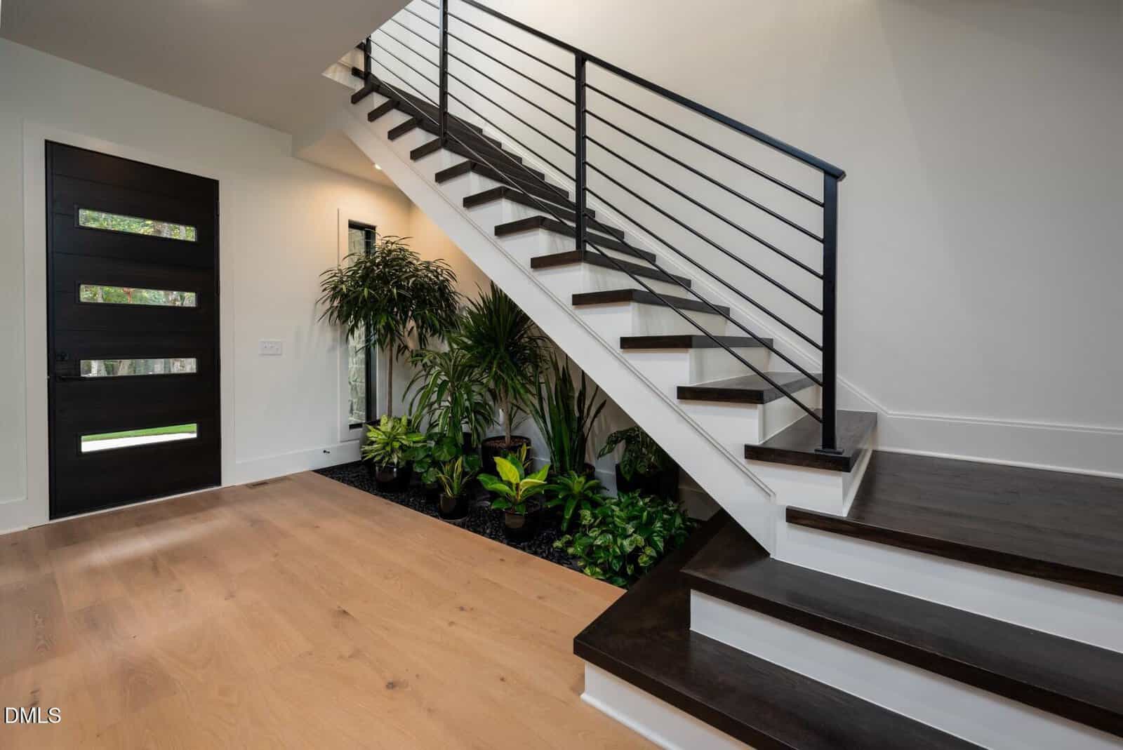A modern entryway with light wood flooring, a dark wooden front door, and a staircase with black railings. Green indoor plants fill the space beneath the stairs, creating a fresh, inviting atmosphere.