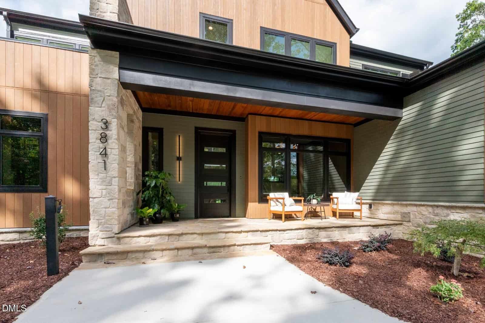 Modern house entrance with wood and stone exterior, black-framed glass door, two chairs with a table on a small porch, and minimal landscaping along a concrete walkway.
