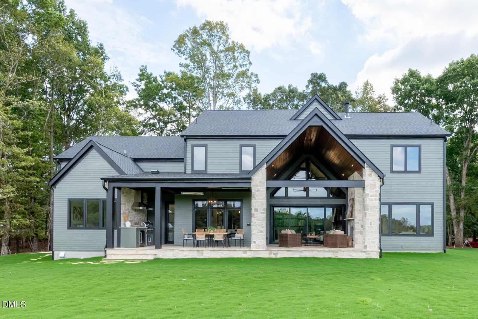 A modern two-story house with gray siding, large windows, and stone accents, featuring a covered patio area with outdoor seating on a grassy lawn, surrounded by trees.