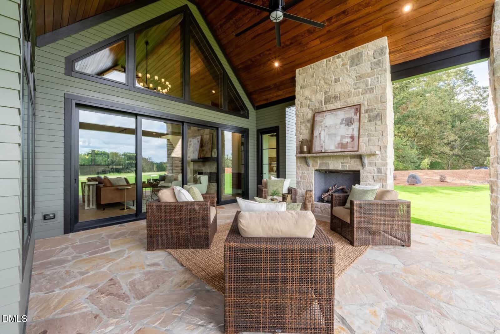 A covered patio features wicker furniture with beige cushions arranged around a stone fireplace. The space has stone flooring, a wood-paneled ceiling, large glass doors, and a view of a grassy outdoor area.
