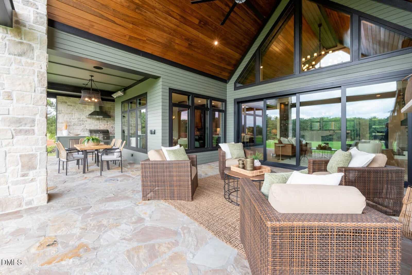 Spacious covered patio with wicker furniture and beige cushions, stone flooring, wood-paneled ceiling, large windows, and a dining area with chairs and a table, overlooking a green outdoor landscape.