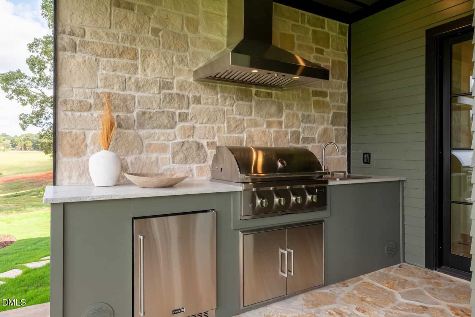 Outdoor kitchen with a stainless steel grill, vent hood, and mini fridge set in a stone and green cabinetry counter. Decorative vase and bowl sit on the countertop. Stone wall and flooring visible. Lawn seen in the background.