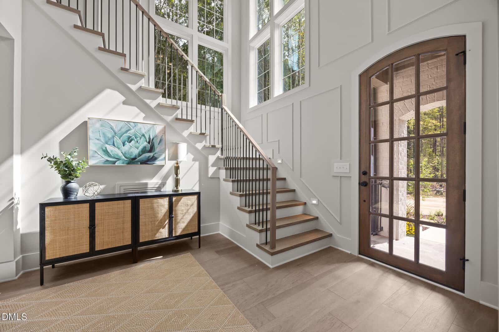 Bright entryway with large windows, wooden stairs with metal railings, a glass-paneled door, a console table with decor and a plant, light-colored walls, and a patterned rug on a hardwood floor.