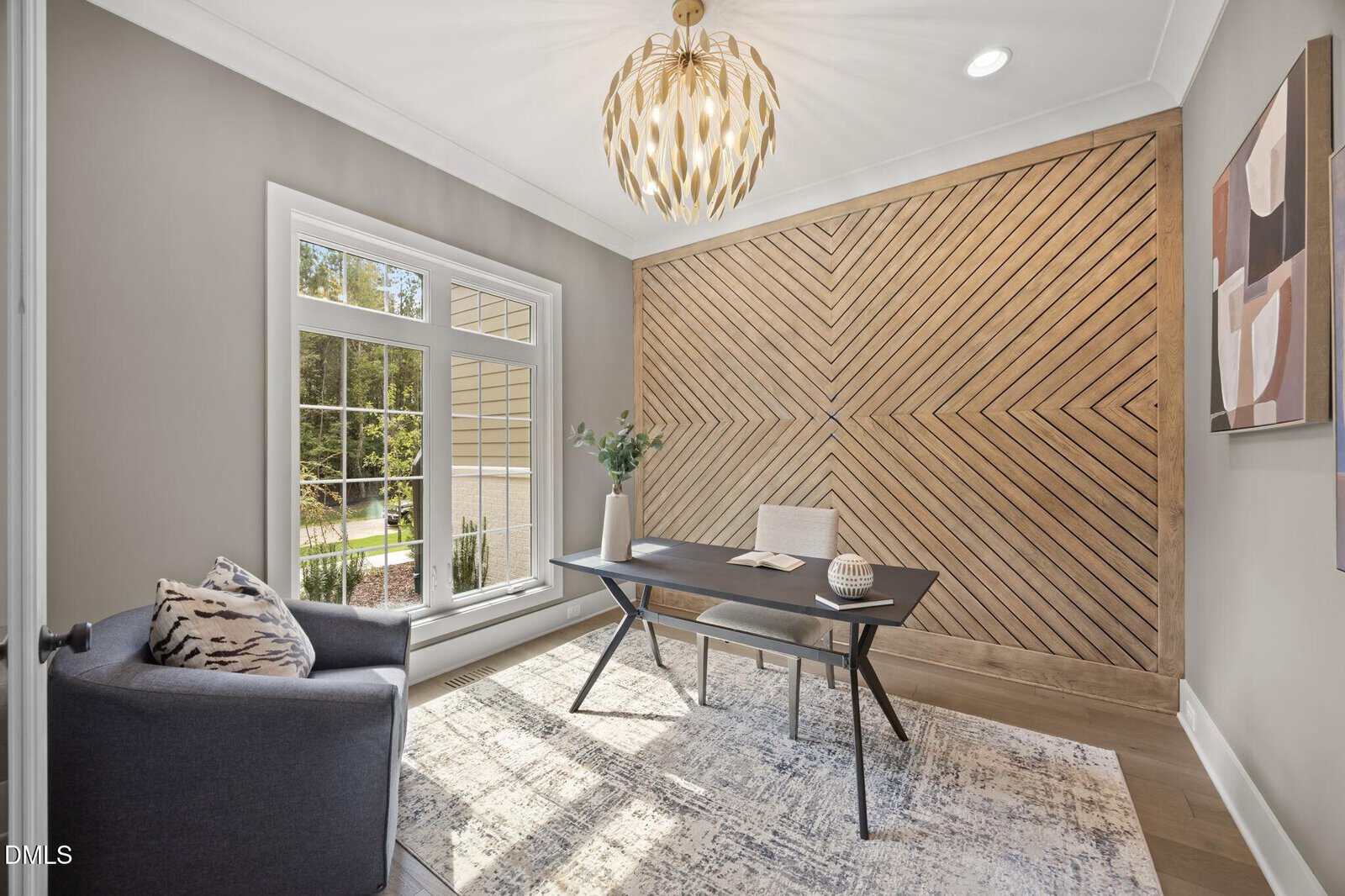 A modern home office with a large window, geometric wood accent wall, black desk, beige chair, gray armchair, abstract wall art, potted plant, and a contemporary chandelier, all in neutral tones.